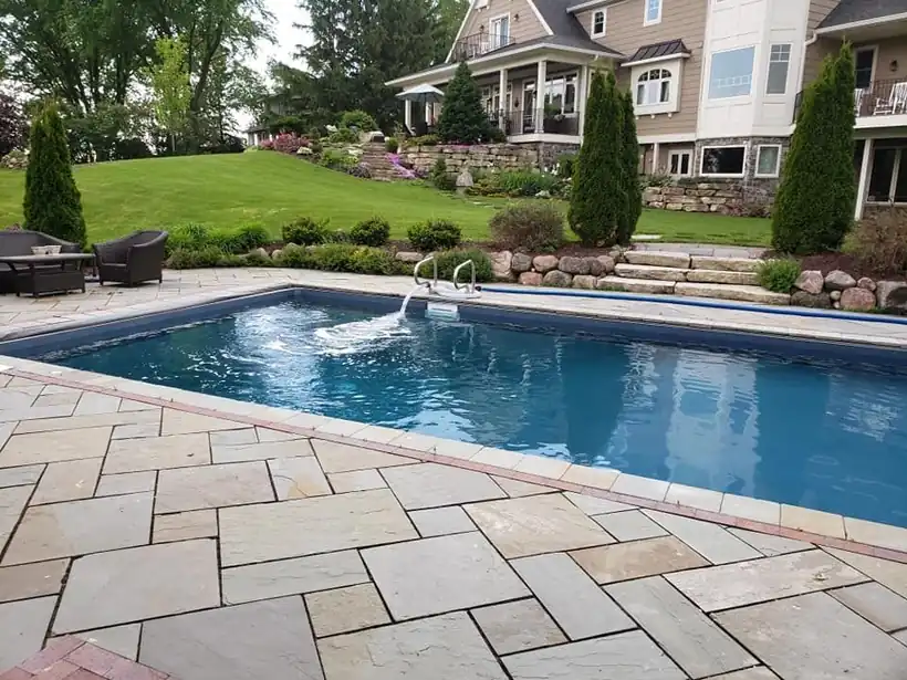A finished rectangular in-ground pool with a stone paver deck below a landscaped hillside home