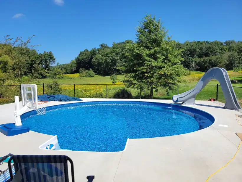 A round pool with a slide and a basketball hoop on a concrete deck, open countryside behind