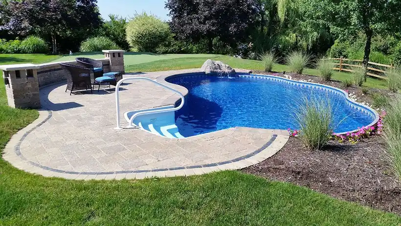 A kidney-shaped in-ground pool with clear blue water beside a paver patio and outdoor kitchen